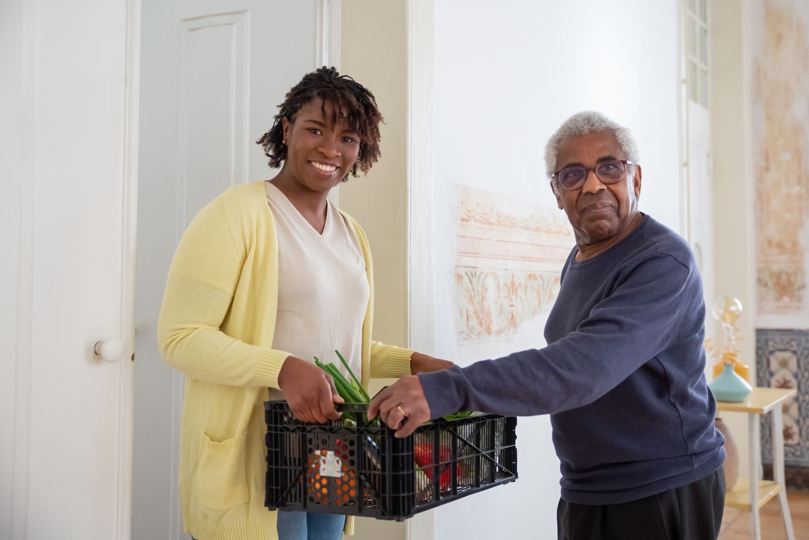 A caregiver bringing a crate of groceries into an older man’s home