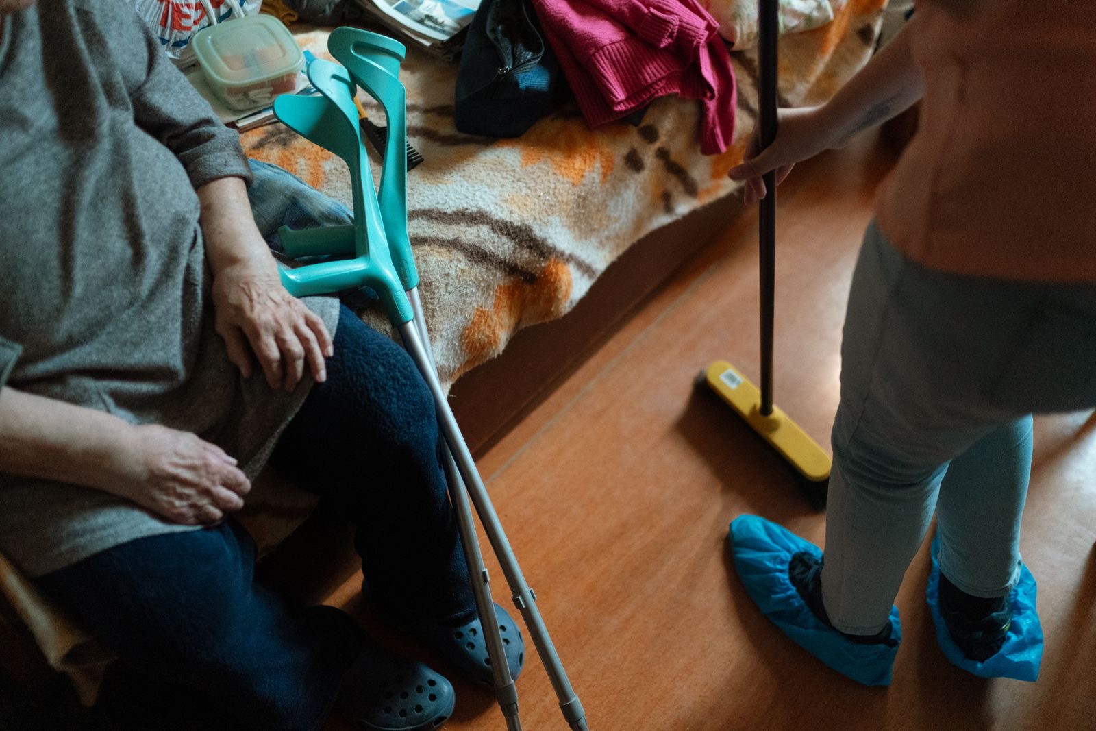 A caregiver sweeping while an older woman with crutches sits nearby