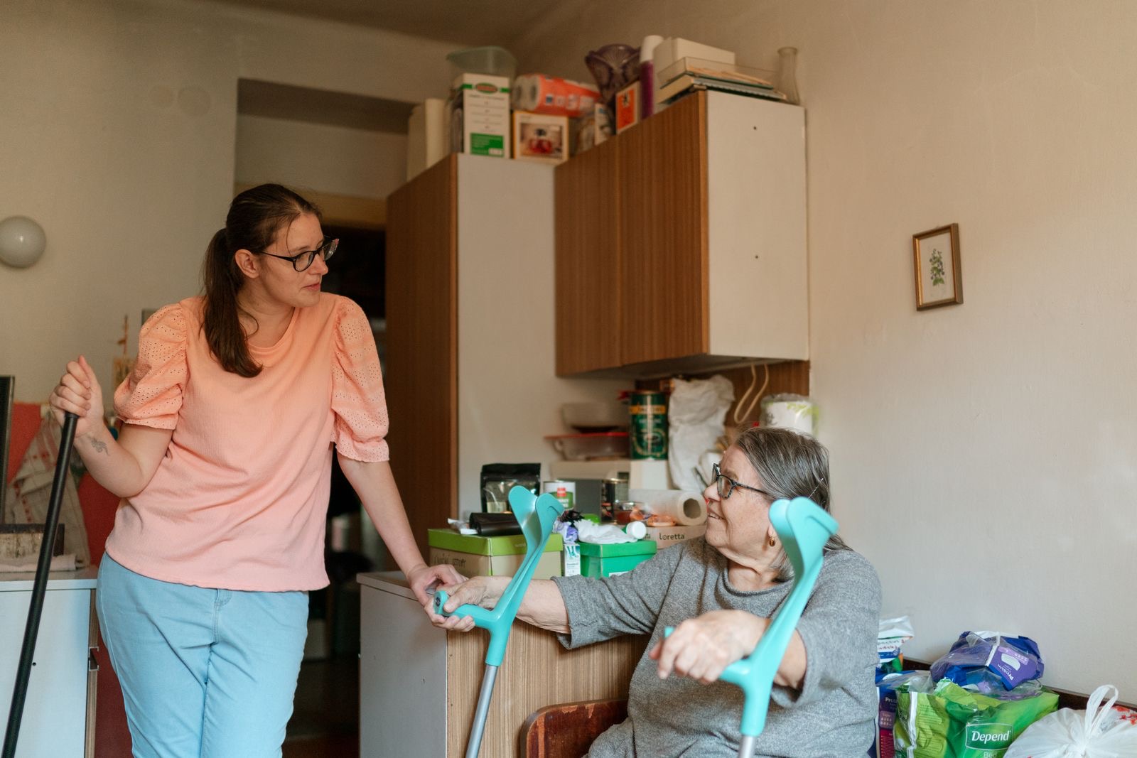 A caregiver with an older woman using crutches in her kitchen